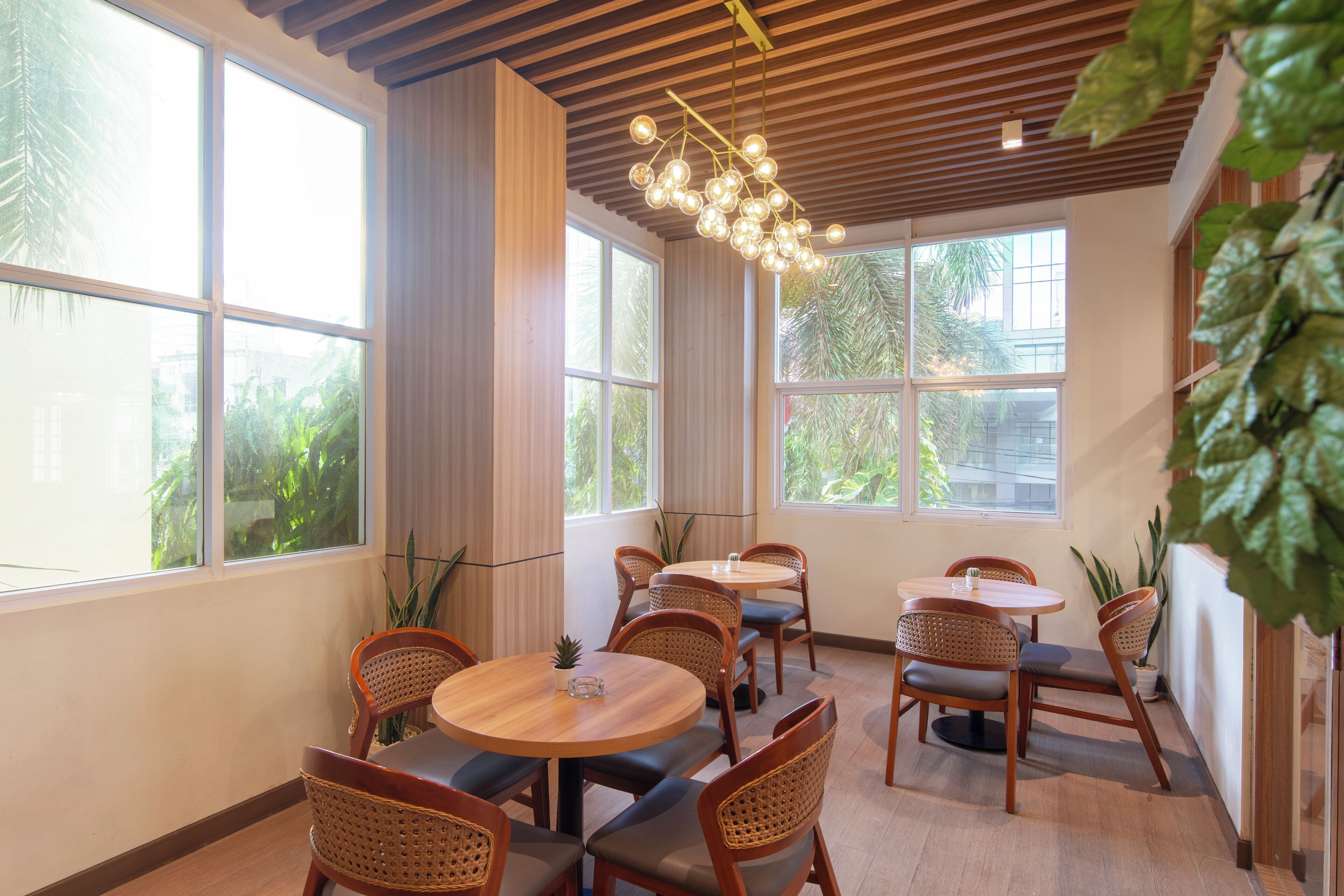 Un comedor con mesas redondas y sillas de mimbre, ventanas amplias con vistas a la vegetación y una lámpara de techo moderna.