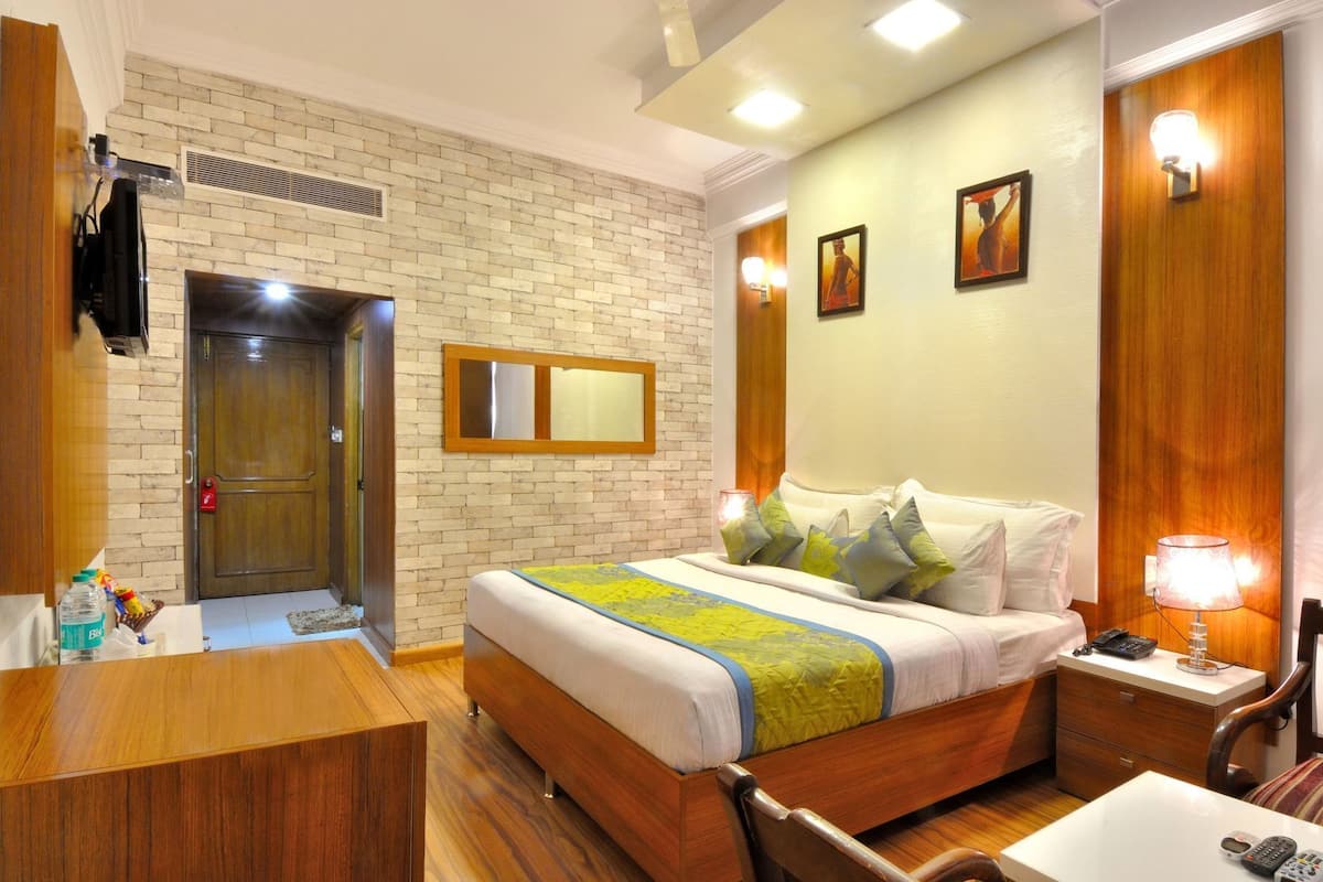 A hotel room with a brick wall, a bed with a green and blue bedspread, a wooden desk, a bedside table with a lamp, and a door with a red handle.