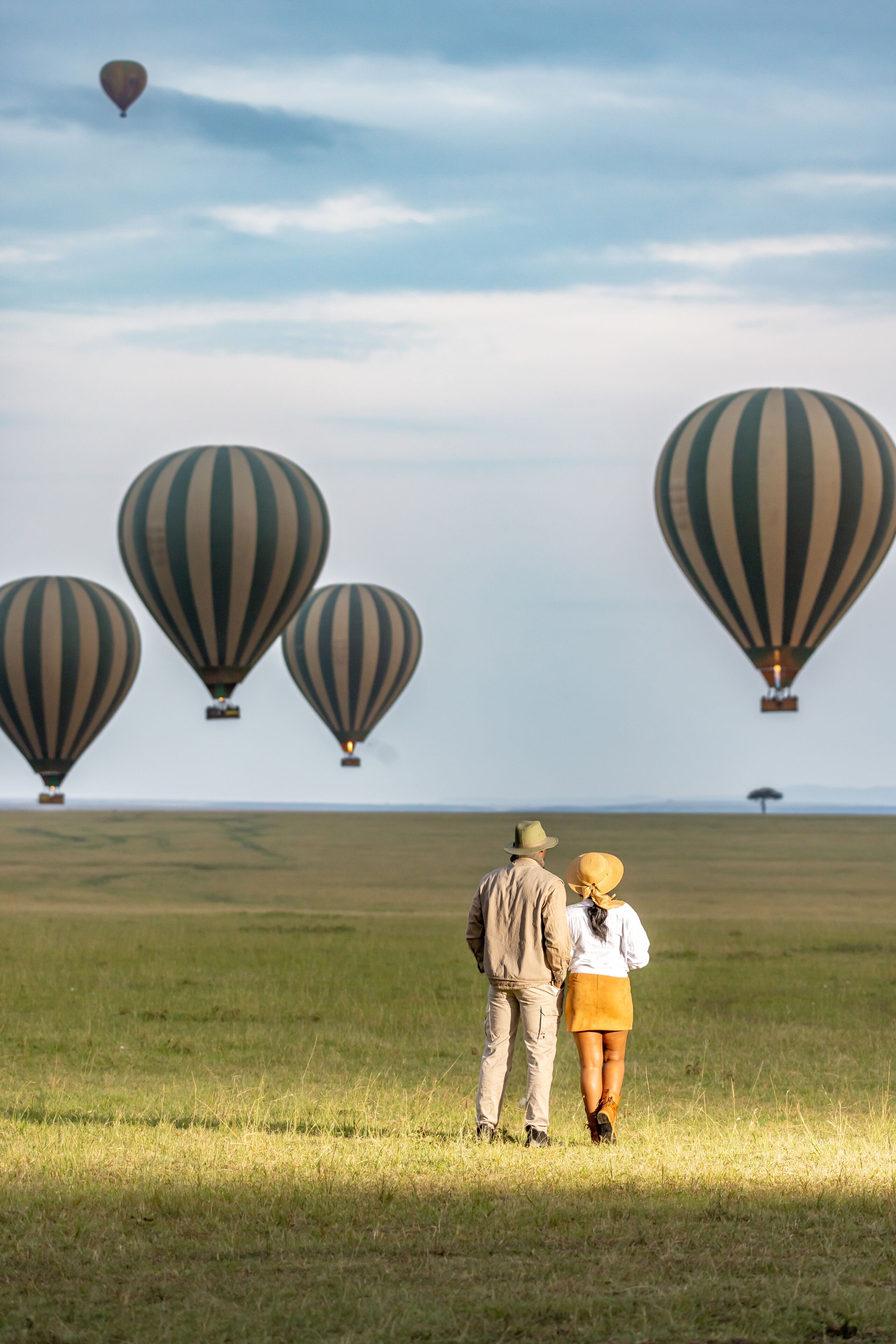 Una pareja de pie en un campo cubierto de hierba, con globos aerostáticos al fondo.