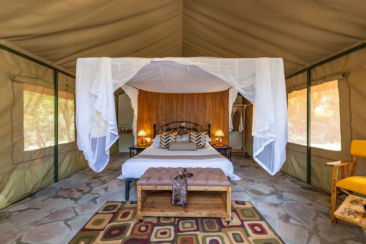 A tented room with a bed, two bedside lamps, a bench, and a chair.