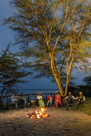 A group of people sitting around a campfire in an outdoor setting with a large tree and a body of water in the background. - Sentrim Elementaita Lodge (Gilgil)