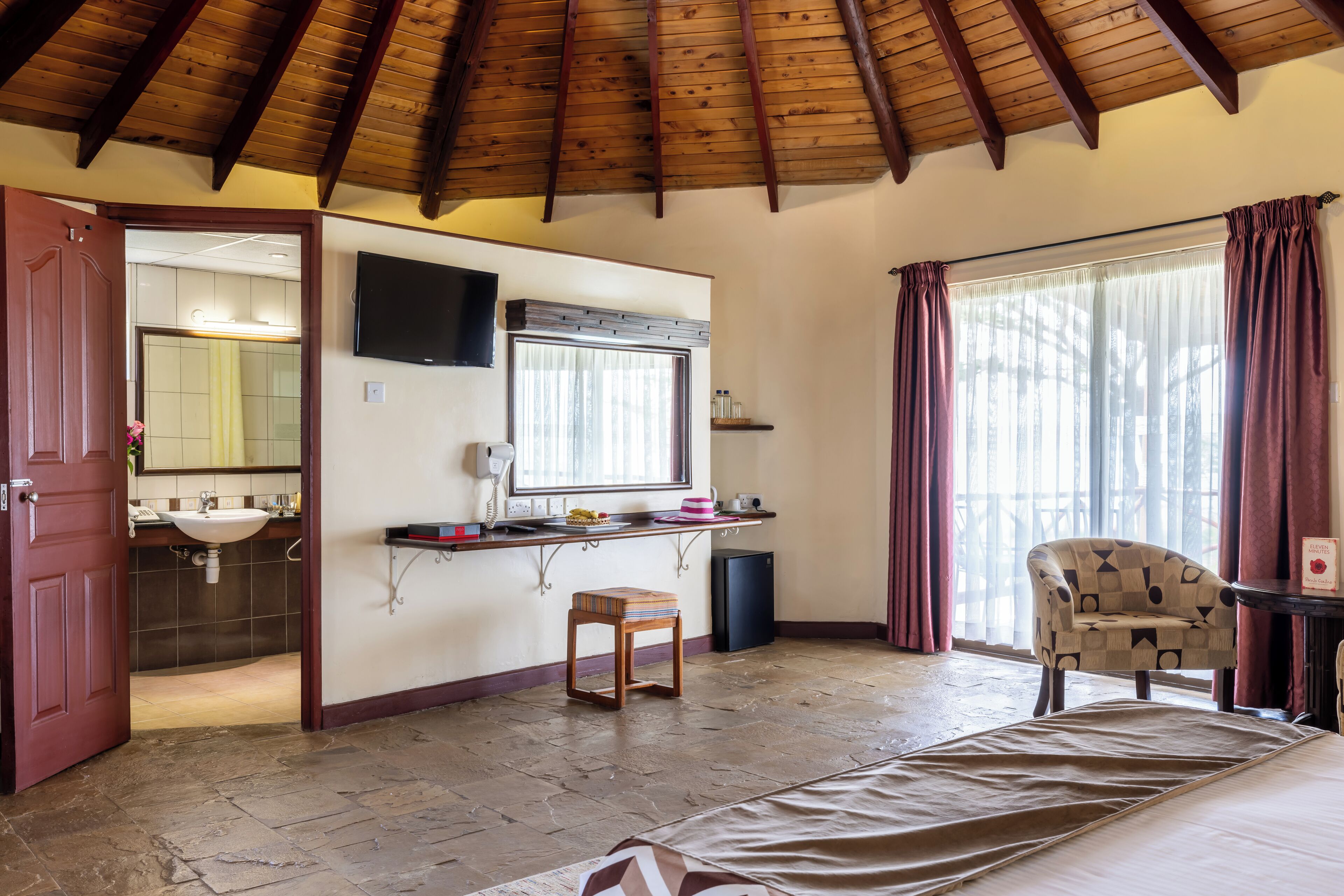 A bedroom with a wooden ceiling, a bed, a chair, a TV, a window with curtains, and a bathroom visible through an open door.
