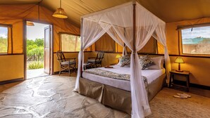 A four-poster canopy bed with a stone floor, a small table with a lamp, and a chair in a tent-style room. - Sentrim Tsavo Lodge (Voi)