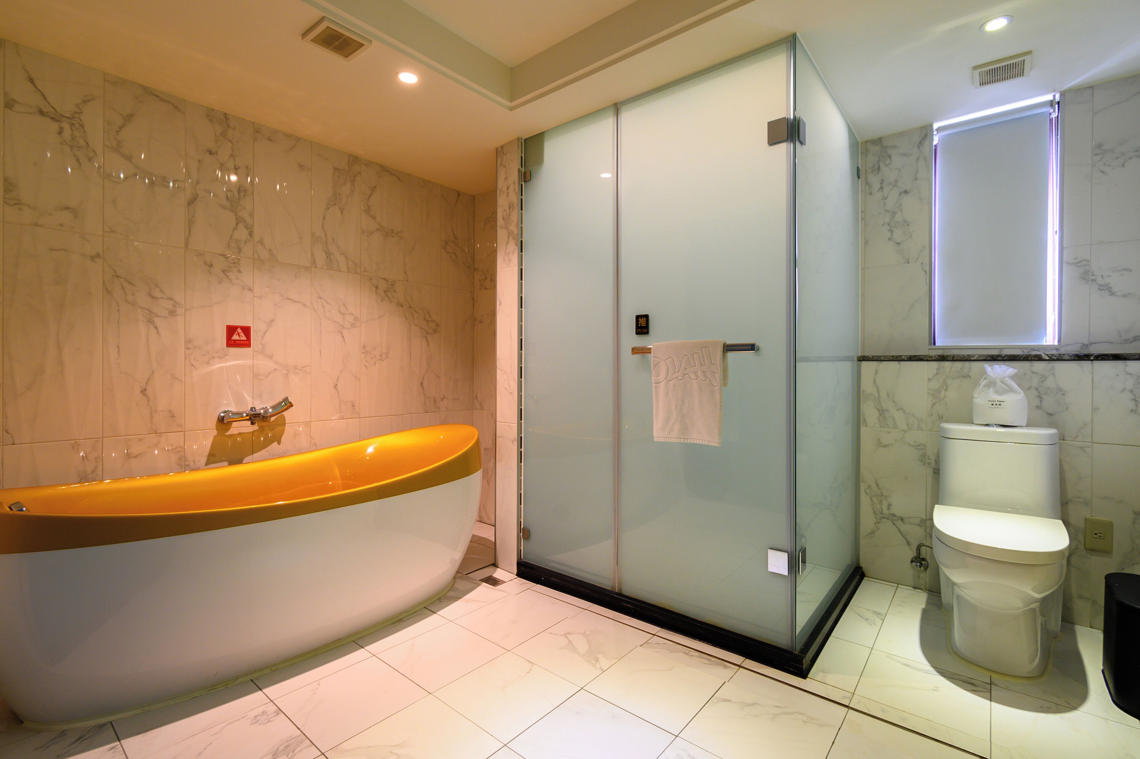 A modern bathroom with a marble wall, a glass-enclosed shower, a white toilet, and a yellow bathtub.