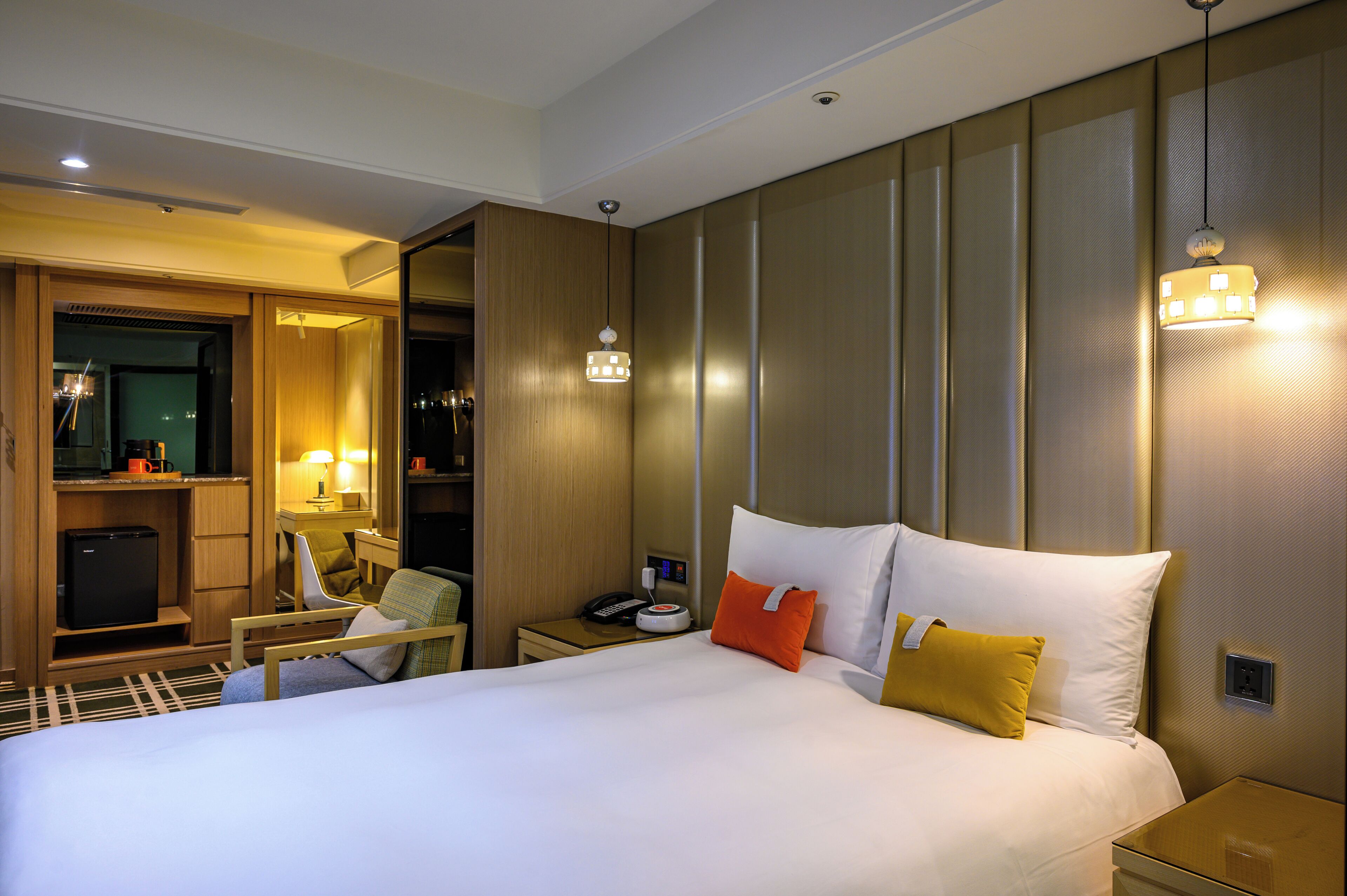 A hotel room with a large bed, bedside tables, a desk with a chair, a wardrobe, and a small kitchenette.