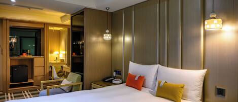 A hotel room with a large bed, bedside tables, a desk with a chair, a wardrobe, and a small kitchenette.