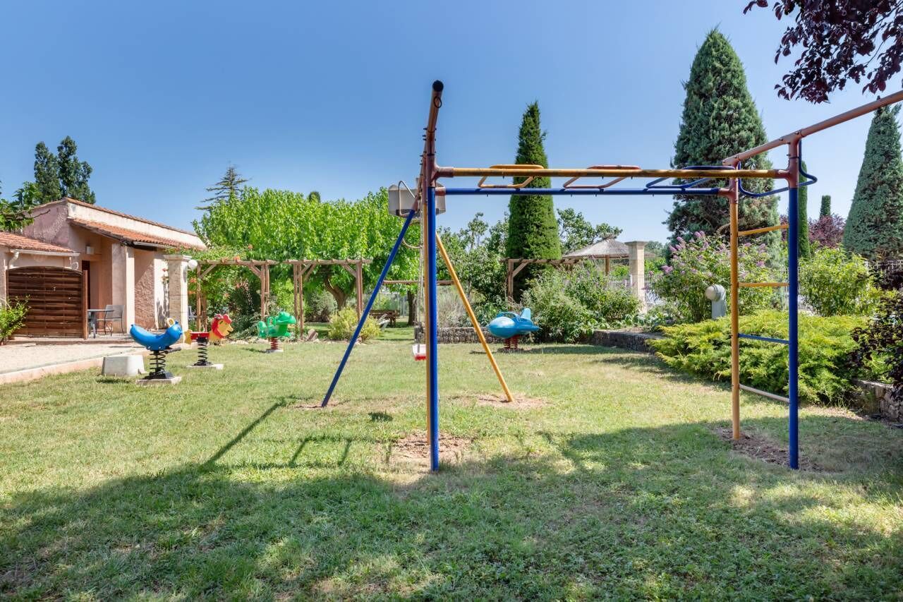 A playground with swings and climbing structures in a grassy area with trees and a building in the background.