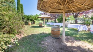A garden with a thatched umbrella, white tables, and barrels.