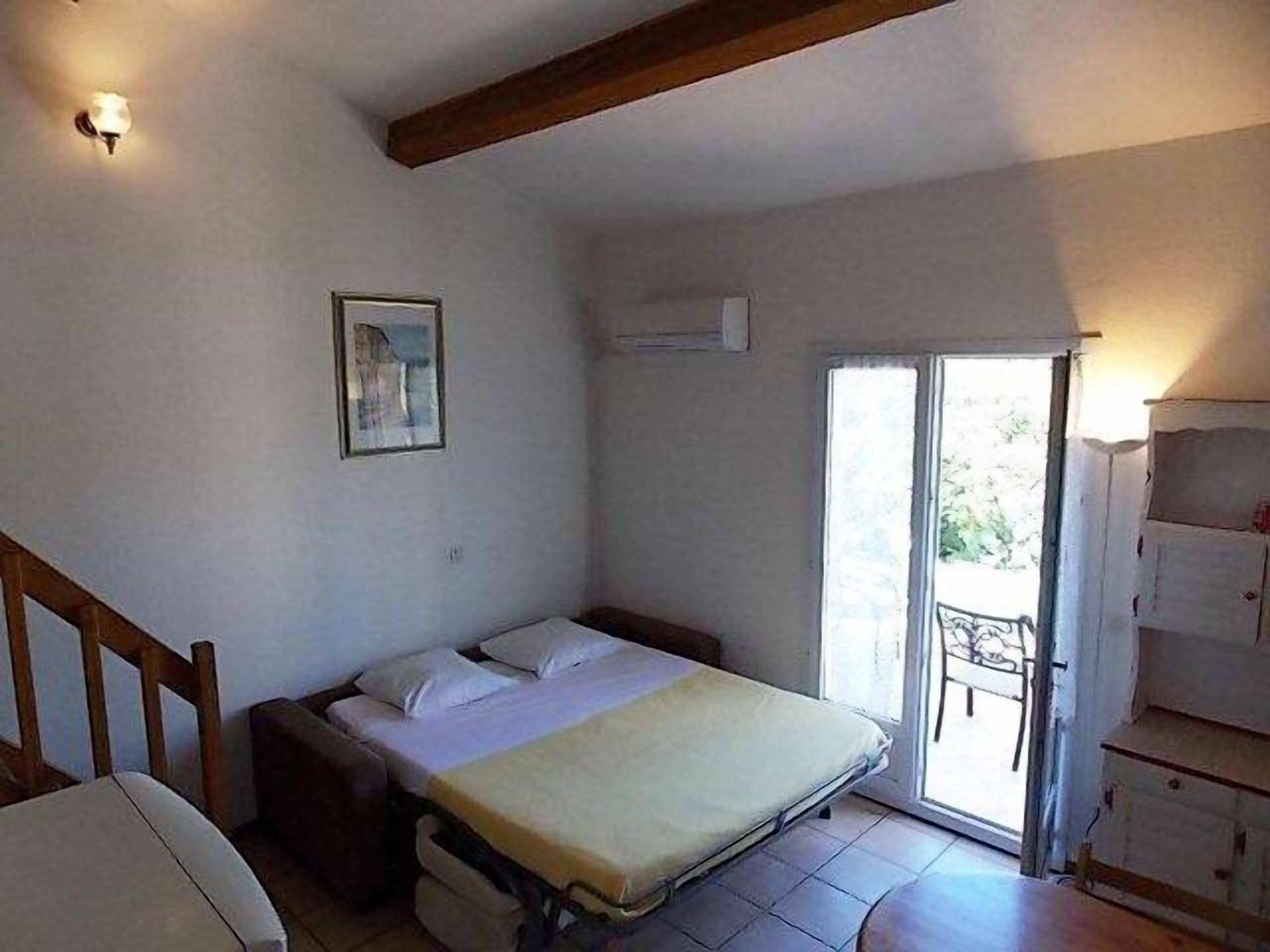 A bedroom with a bed, a chair, a small table, a picture on the wall, and a sliding door leading to an outdoor area.