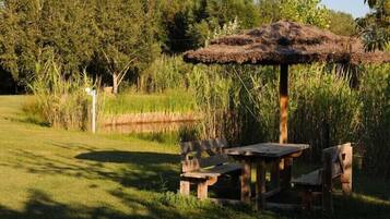 A rustic outdoor seating area with a thatched roof, surrounded by tall grasses and trees.