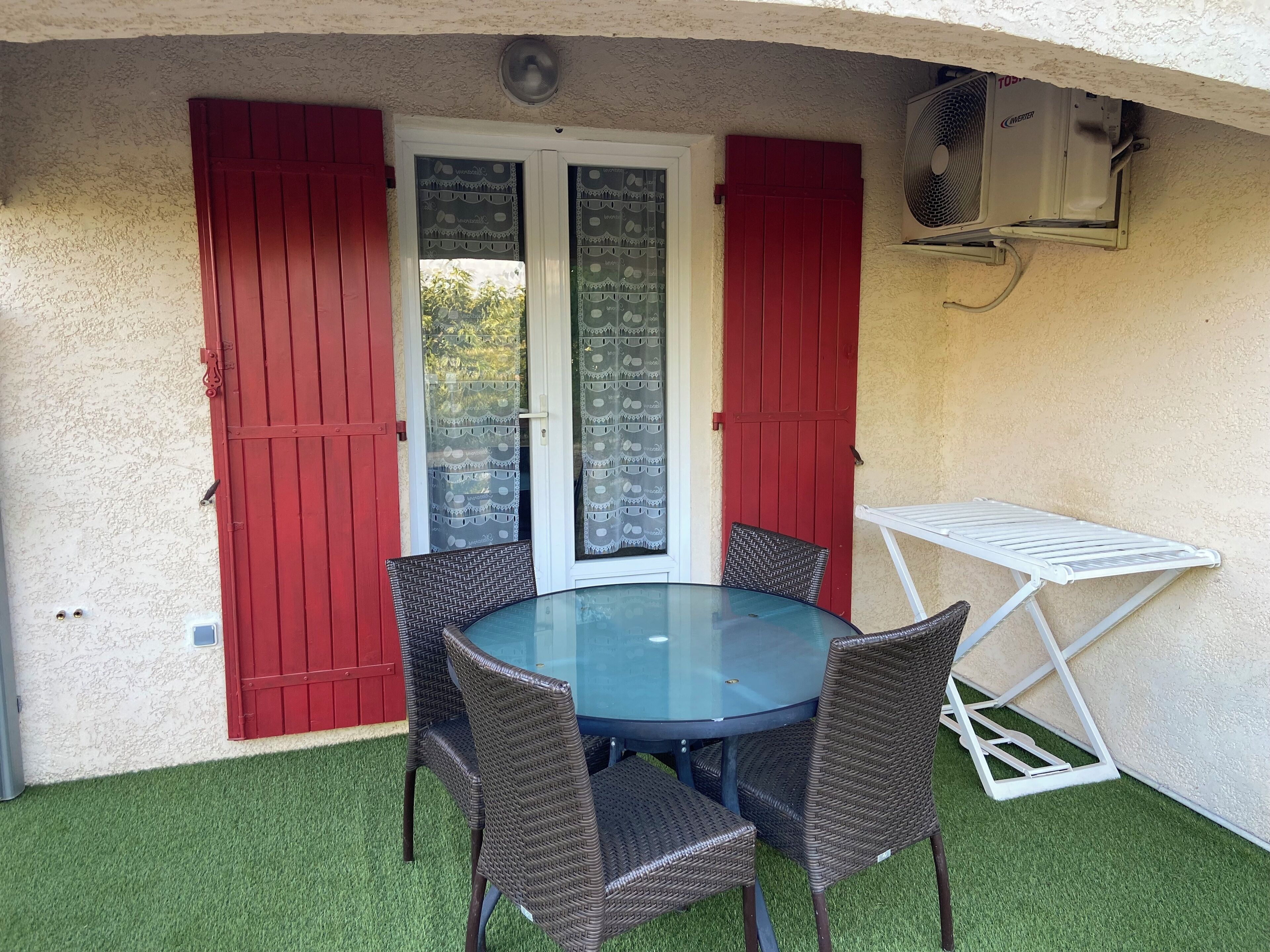 Une terrasse avec une table ronde en verre et quatre chaises en osier, des volets rouges et un étendoir à linge blanc.