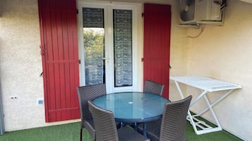 Une terrasse avec une table ronde en verre et quatre chaises en osier, des volets rouges et un étendoir à linge blanc.