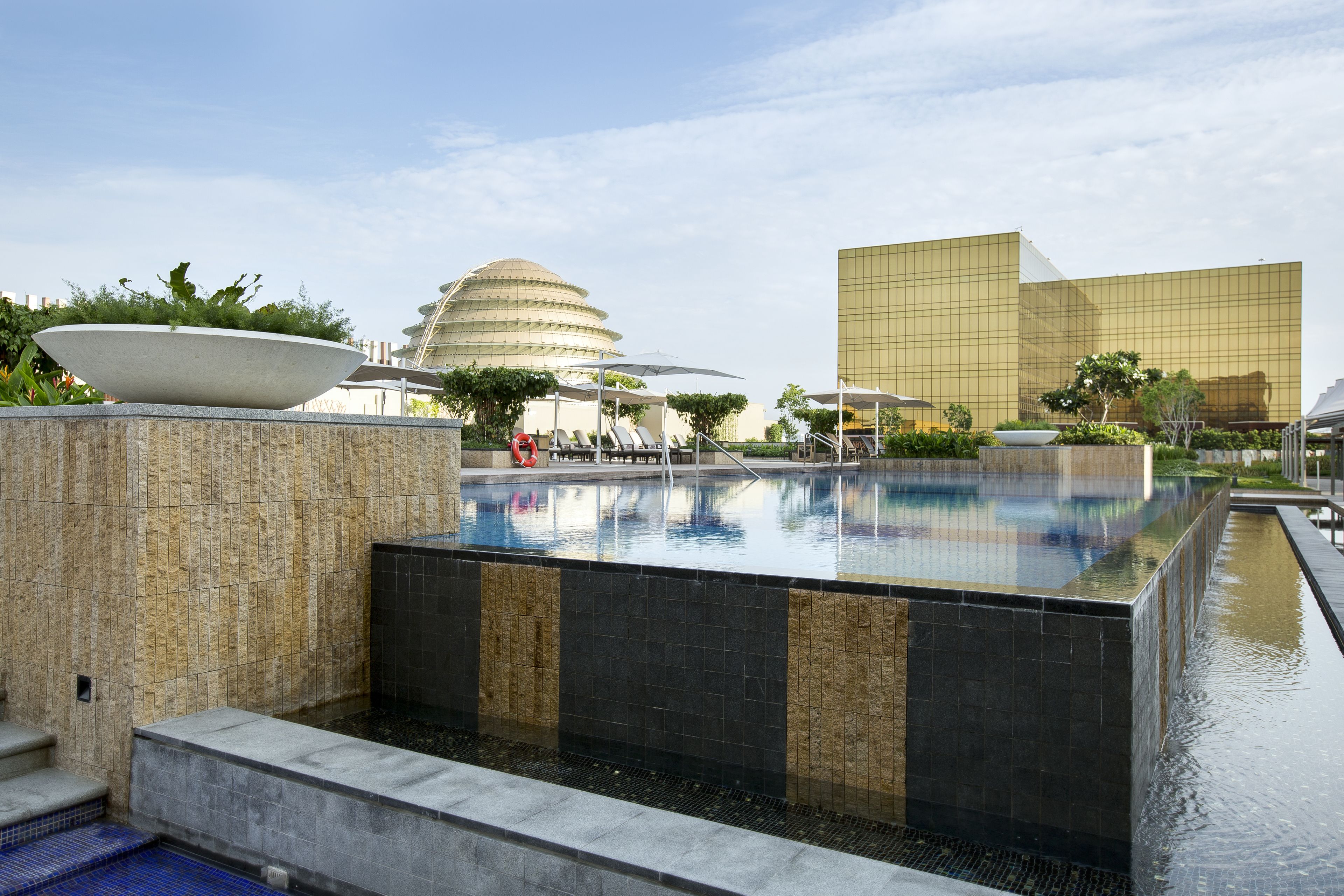 Zona de piscina moderna de hotel con una piscina grande central, una piscina más pequeña adyacente y un área de descanso con sombrillas.