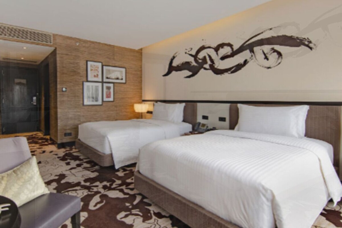Ein Hotelzimmer mit zwei Betten, einem Schreibtisch, einem Stuhl, einer Lampe und einer Wand mit Kunstwerken.