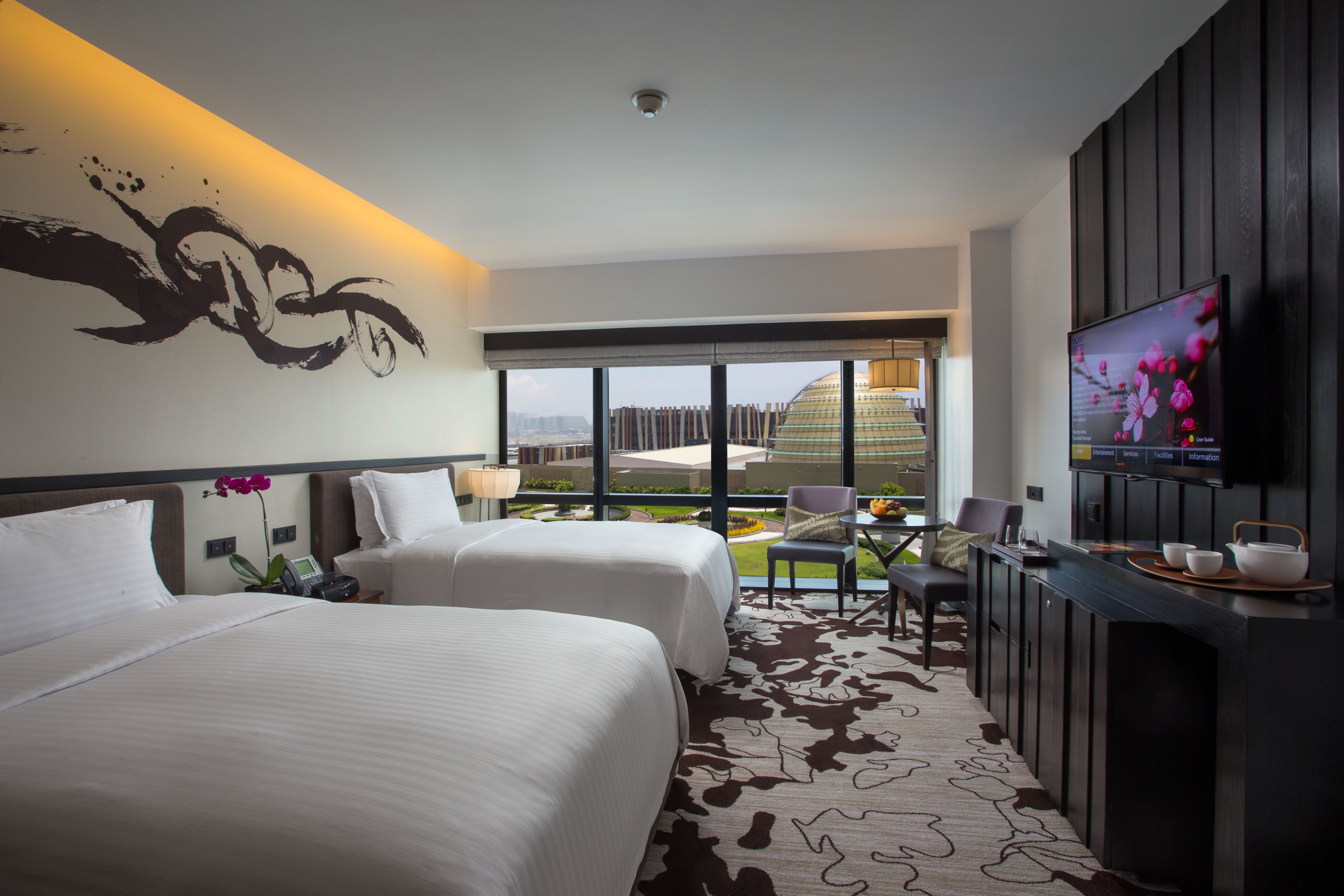 Une chambre d’hôtel avec deux lits, une télévision, un bureau avec une chaise et une grande fenêtre donnant sur la ville.