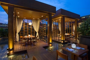 Un área de comedor al aire libre con mesas y sillas de madera, una piscina y vistas del horizonte de la ciudad al anochecer.