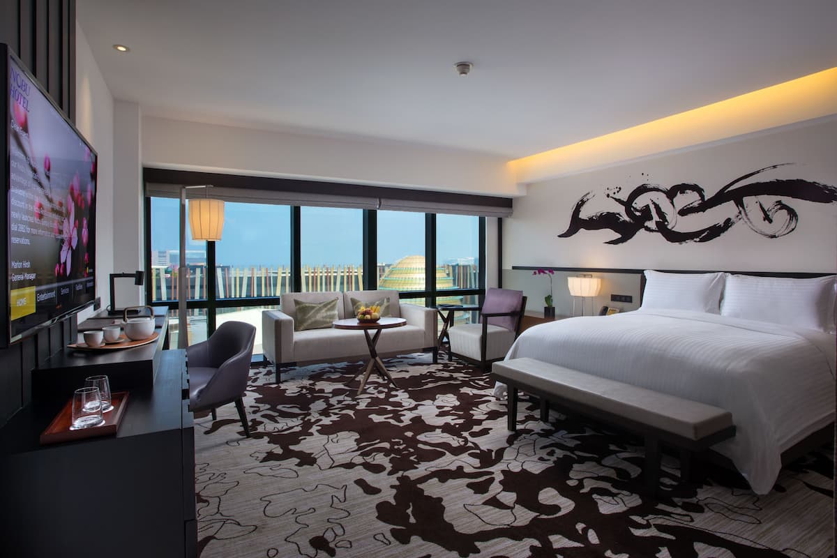Una habitación de hotel moderna con una cama grande, un sofá, un escritorio con televisor y vistas a la ciudad a través de amplias ventanas.