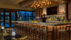 Uma área de bar com um bartender, banquetas, um balcão com diversos tipos de bebidas e vista através de grandes janelas.