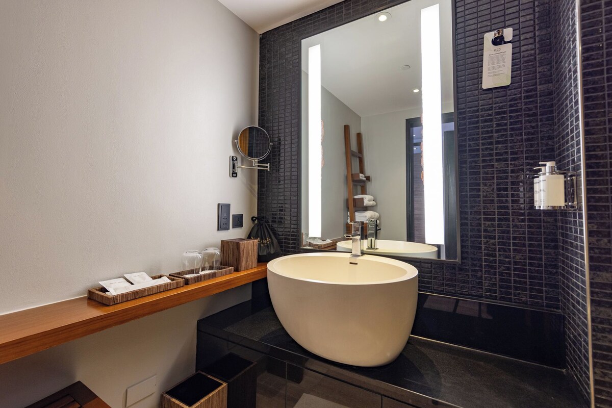 Un baño moderno con un lavamanos amplio, un espejo y un estante de madera con artículos de aseo.