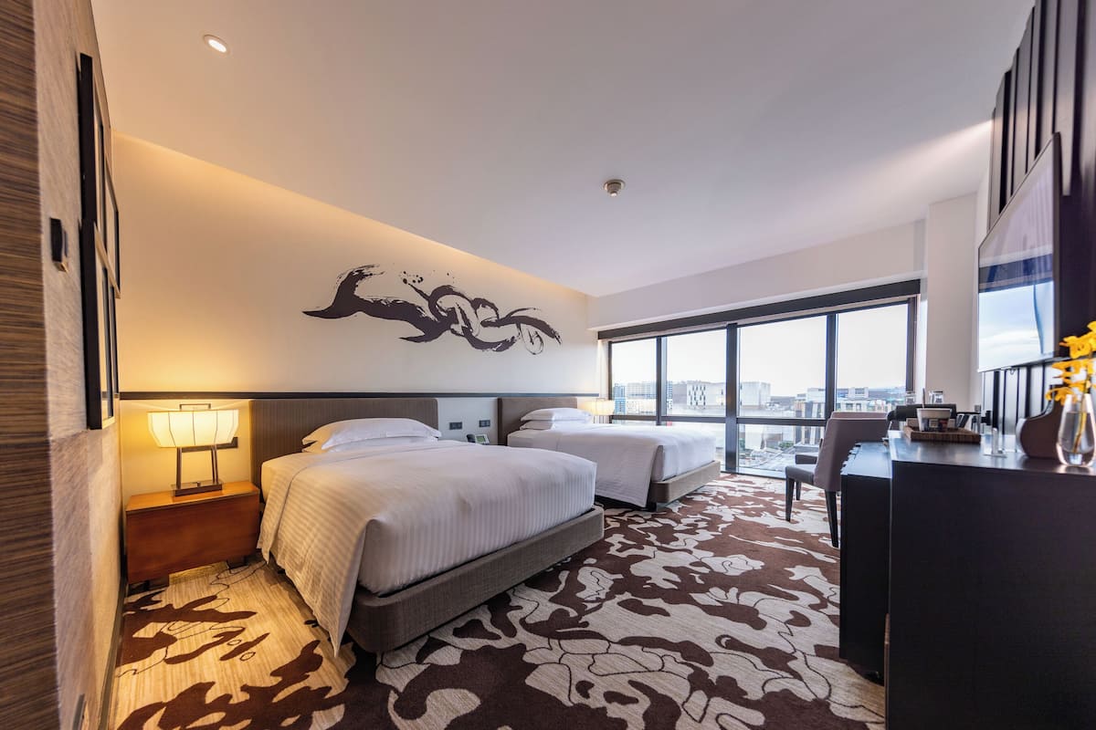 Una habitación de hotel moderna con dos camas, un escritorio y un ventanal amplio con vistas a la ciudad.
