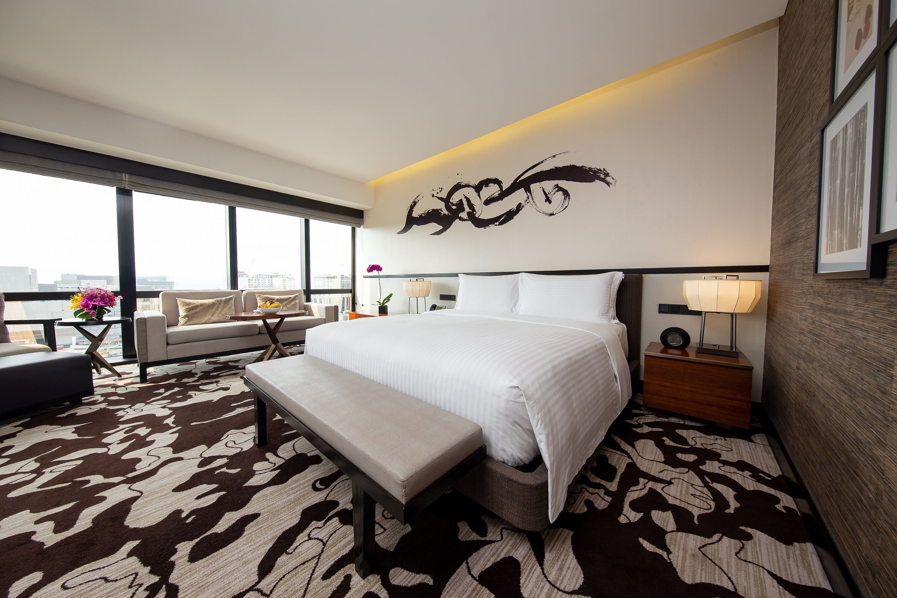 A modern hotel room with a large bed, a bench, a sofa, and a desk. There is a large window providing a view of the city.