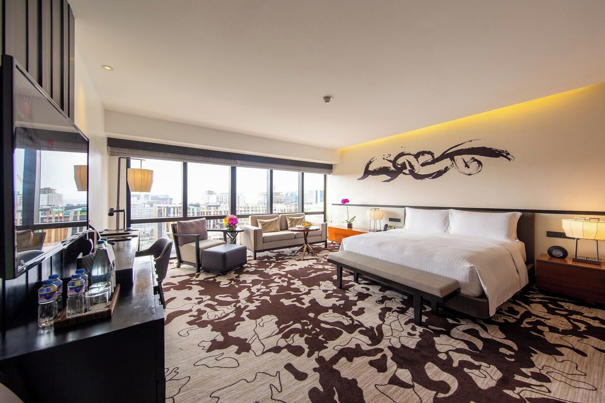 Ein Hotelzimmer mit einem großen Bett, einer Couch, einem Schreibtisch mit Stuhl und Blick auf die Stadt durch große Fenster.
