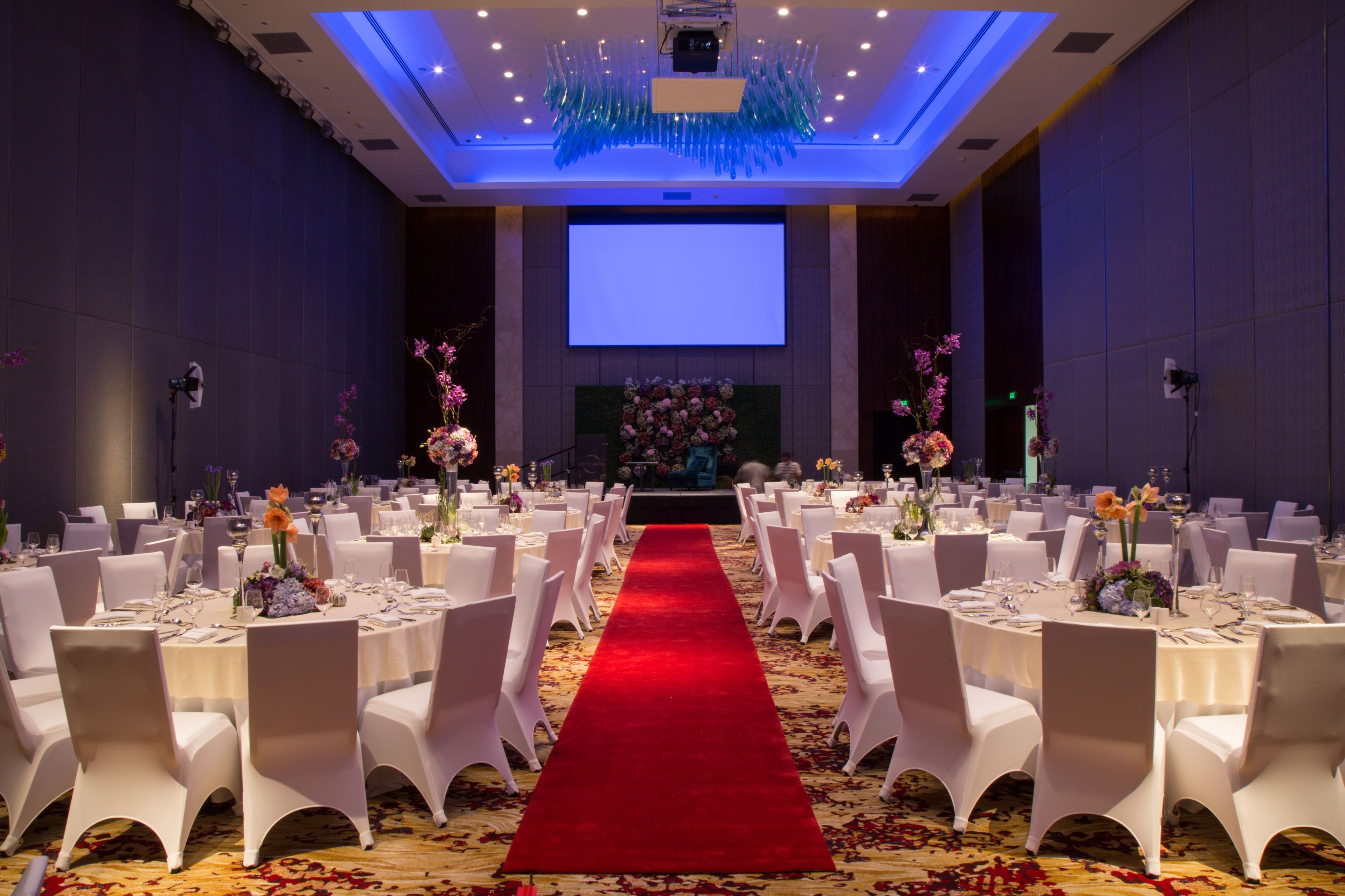A banquet hall with round tables, white chairs, a red carpet, and floral centerpieces.