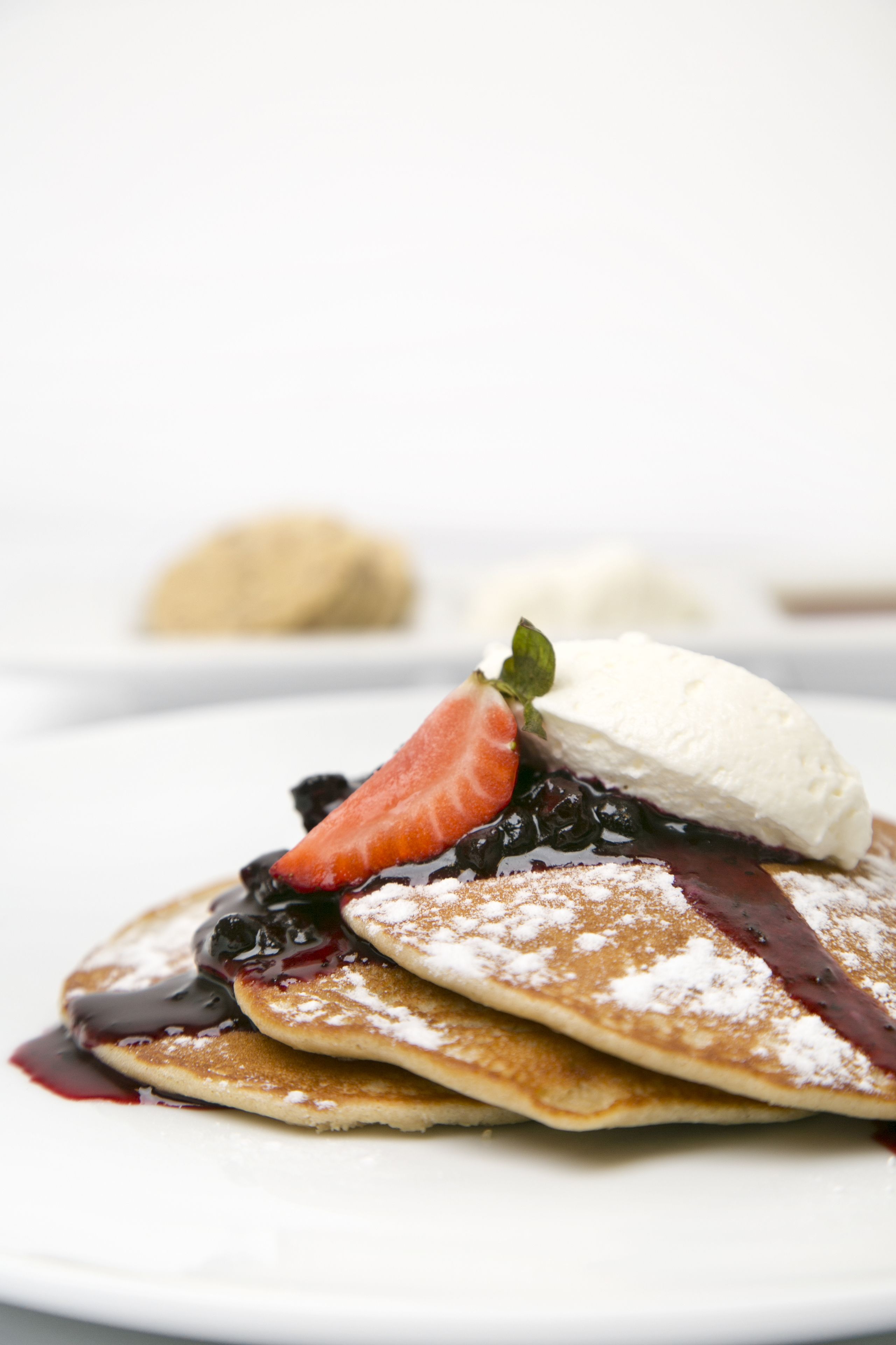 Una pila de tortitas con compota de fresa y arándanos, una cucharada de crema batida y azúcar glas sobre un plato blanco.
