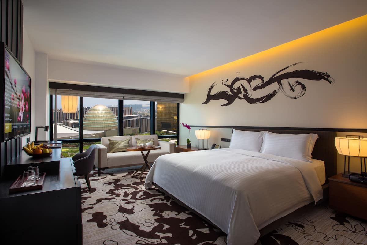 Habitación de hotel moderna con una cama grande, un escritorio con frutero, un sofá, vista a la ciudad y una obra de arte en la pared.