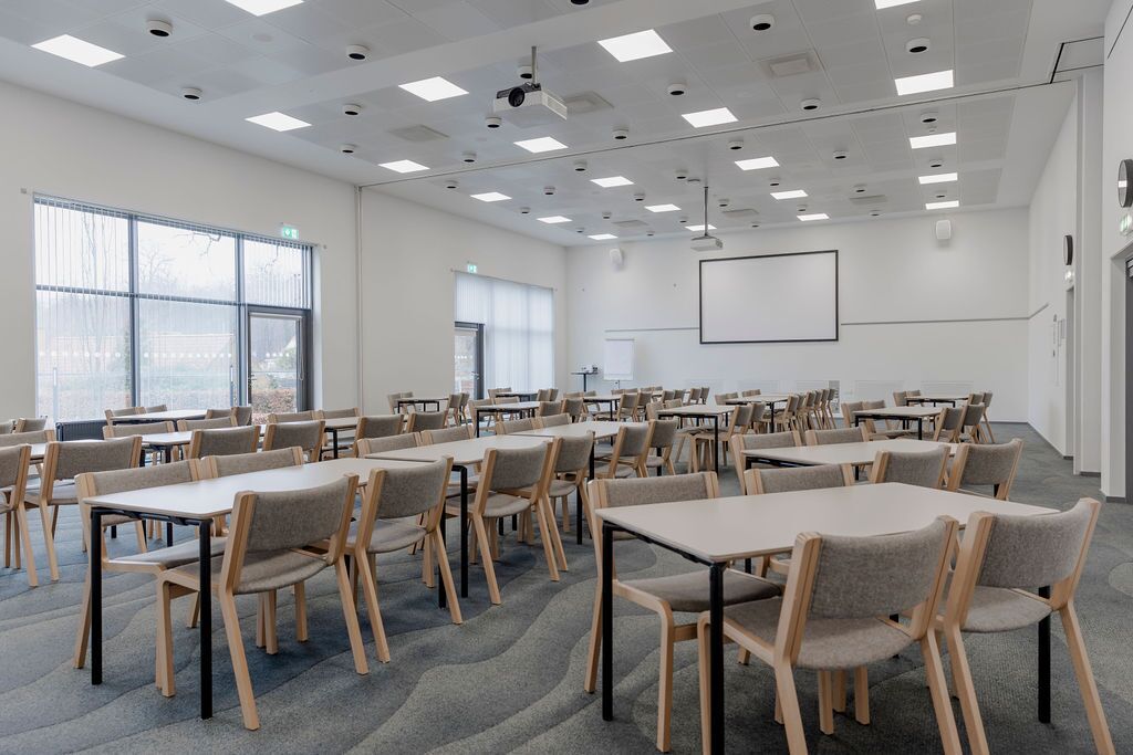 Un salón de conferencias con varias mesas y sillas, ventanas amplias y una pantalla para proyector.