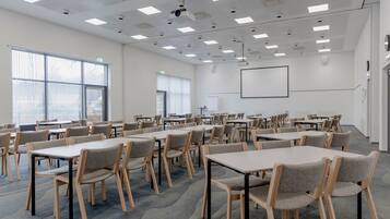 Un salón de conferencias con varias mesas y sillas, ventanas amplias y una pantalla para proyector.