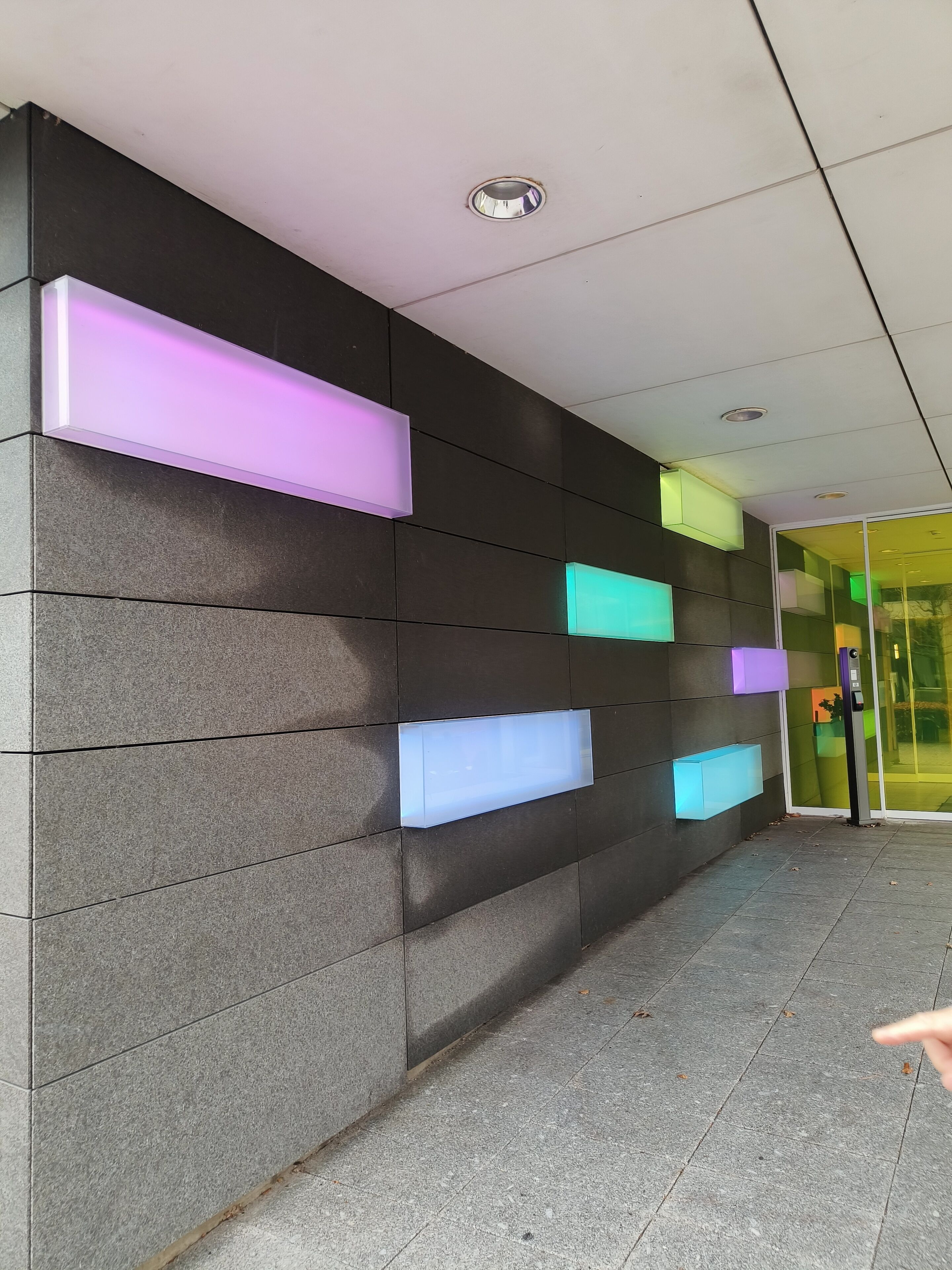 Fachada moderna de un edificio con suelo de baldosas y una pared con paneles rectangulares de colores variados.