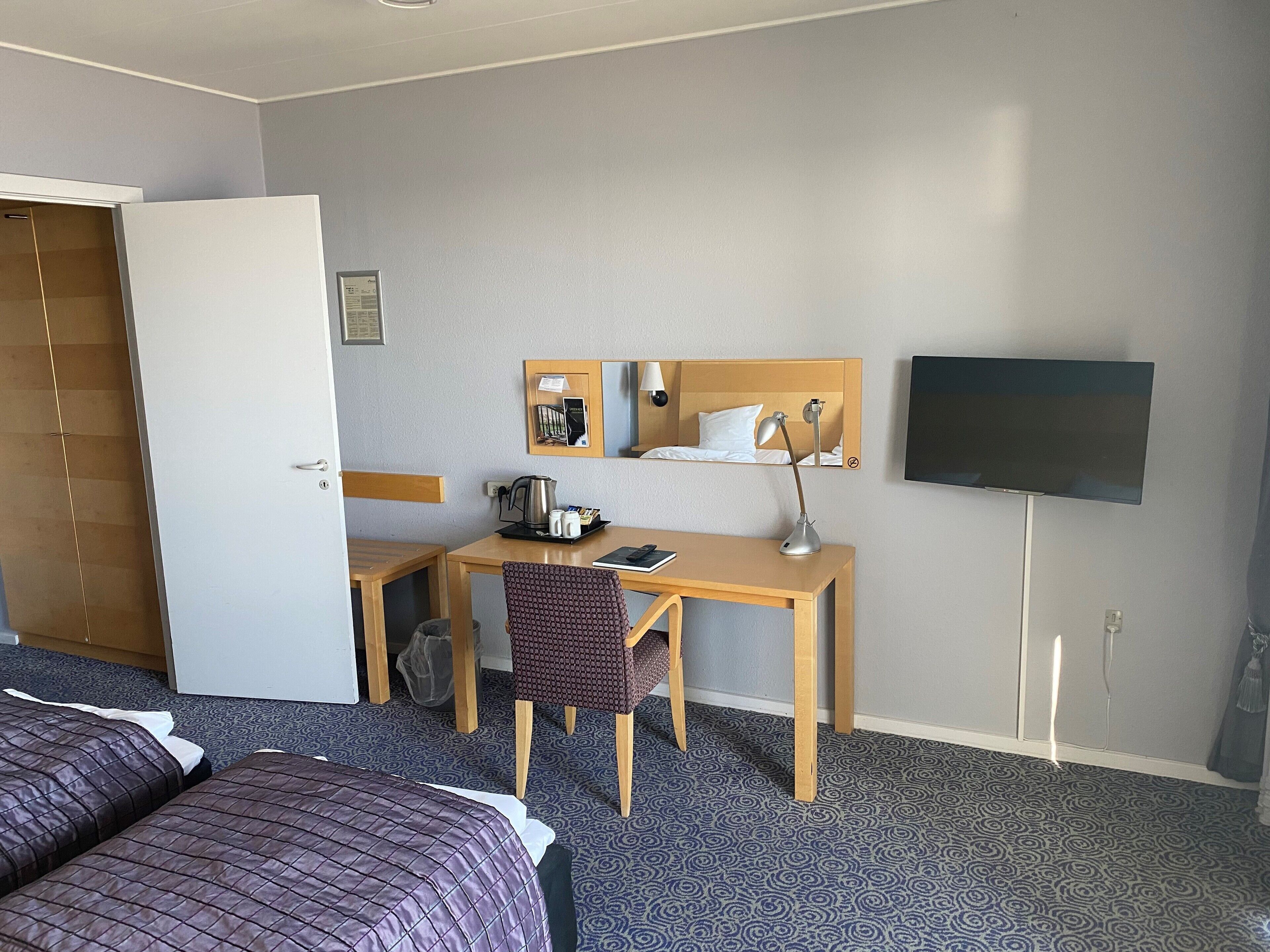 Habitación de hotel con cama, escritorio, silla, televisor y una mesita con tetera y tazas.