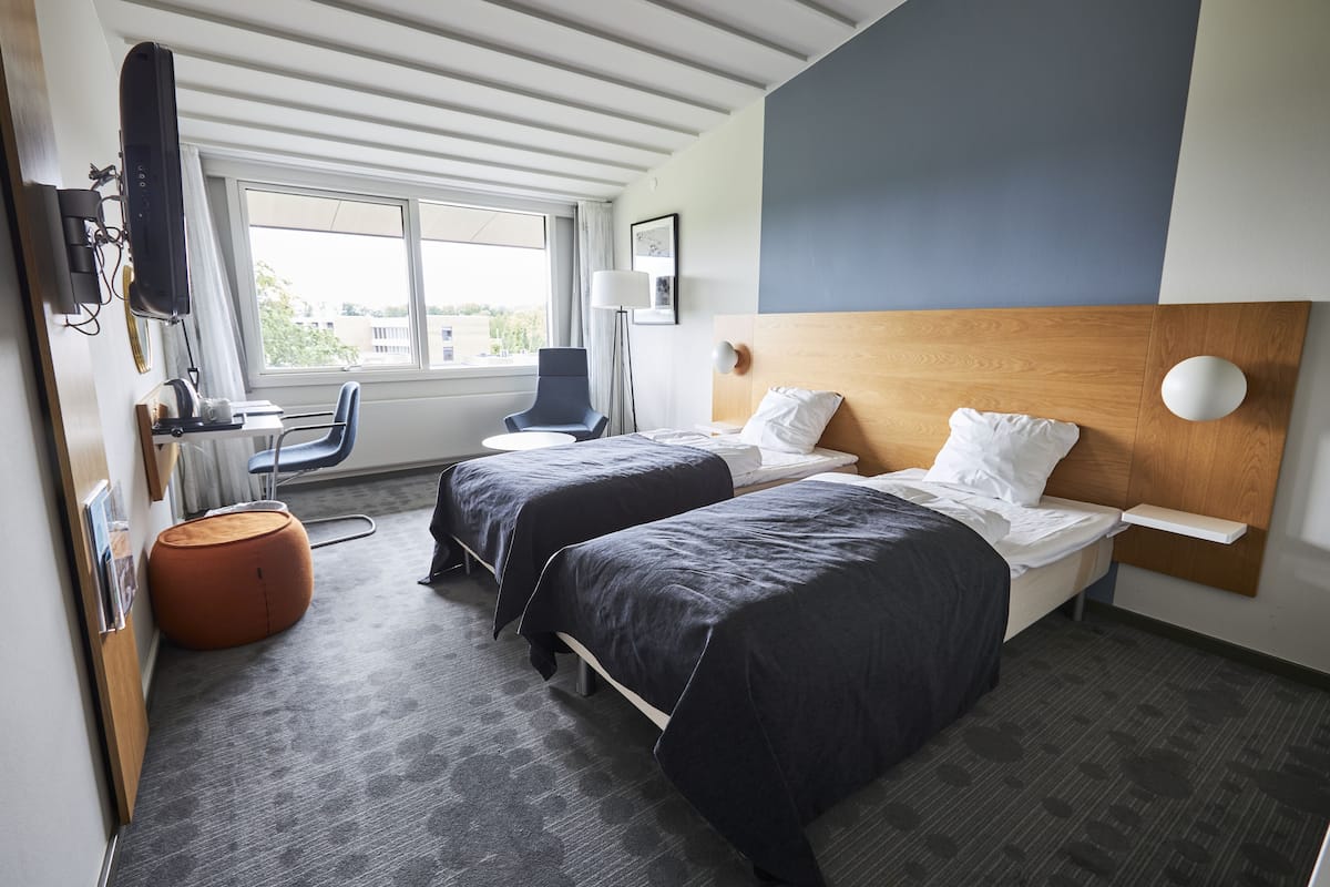 A hotel room with two beds, a desk, a chair, a round ottoman, and a TV.