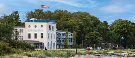 Hotel 'Hotel Nyborg Strand' con una bandera, rodeado de vegetación y rocas.