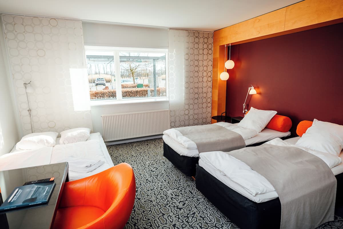 A hotel room with two beds, a desk, an orange chair, and a window with a view of the street.