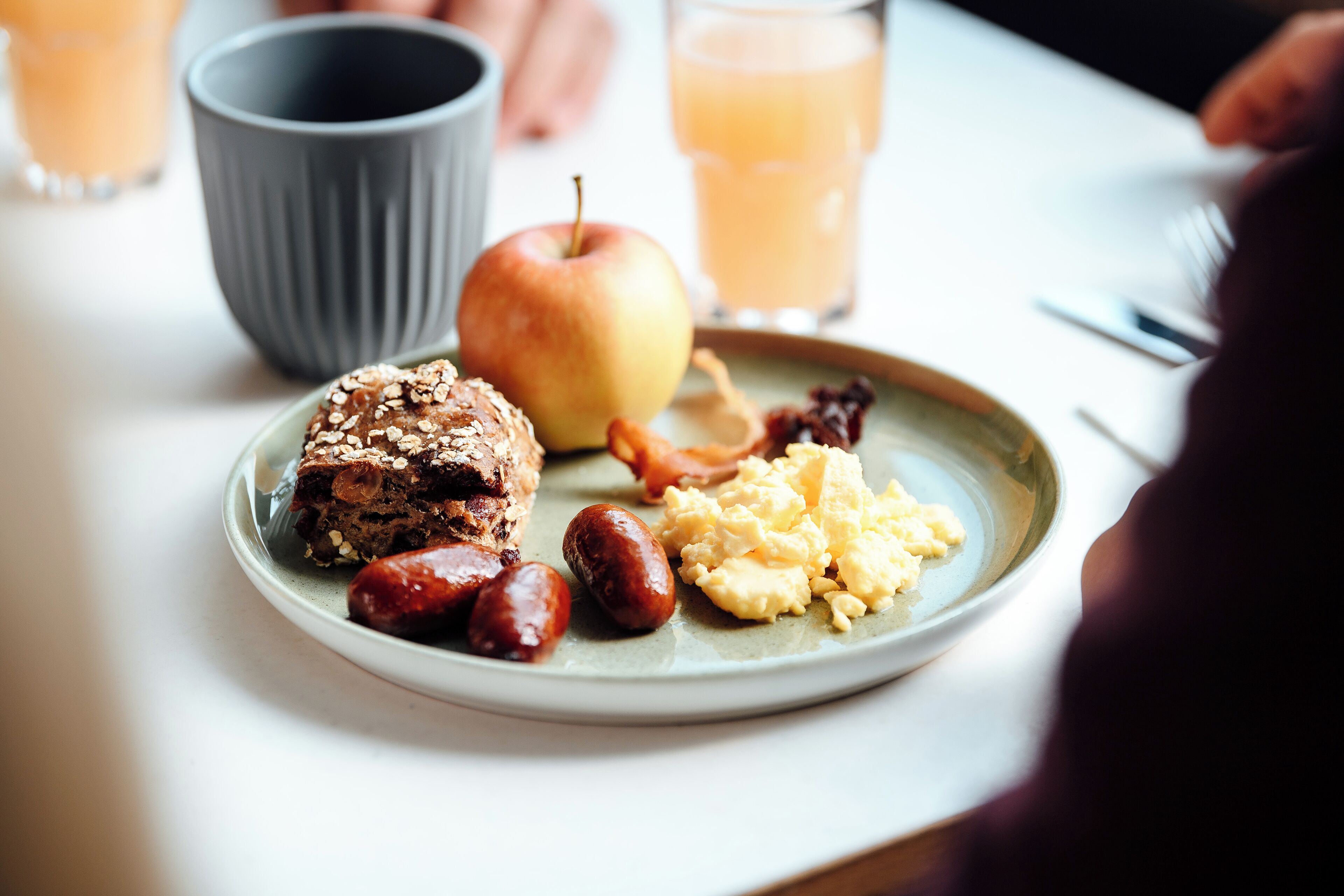 A plate with a slice of cake, an apple, scrambled eggs, and sausages.