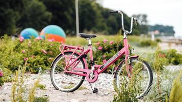 A pink bicycle with a basket and a chain guard, parked on a gravel path with greenery and flowers in the background.