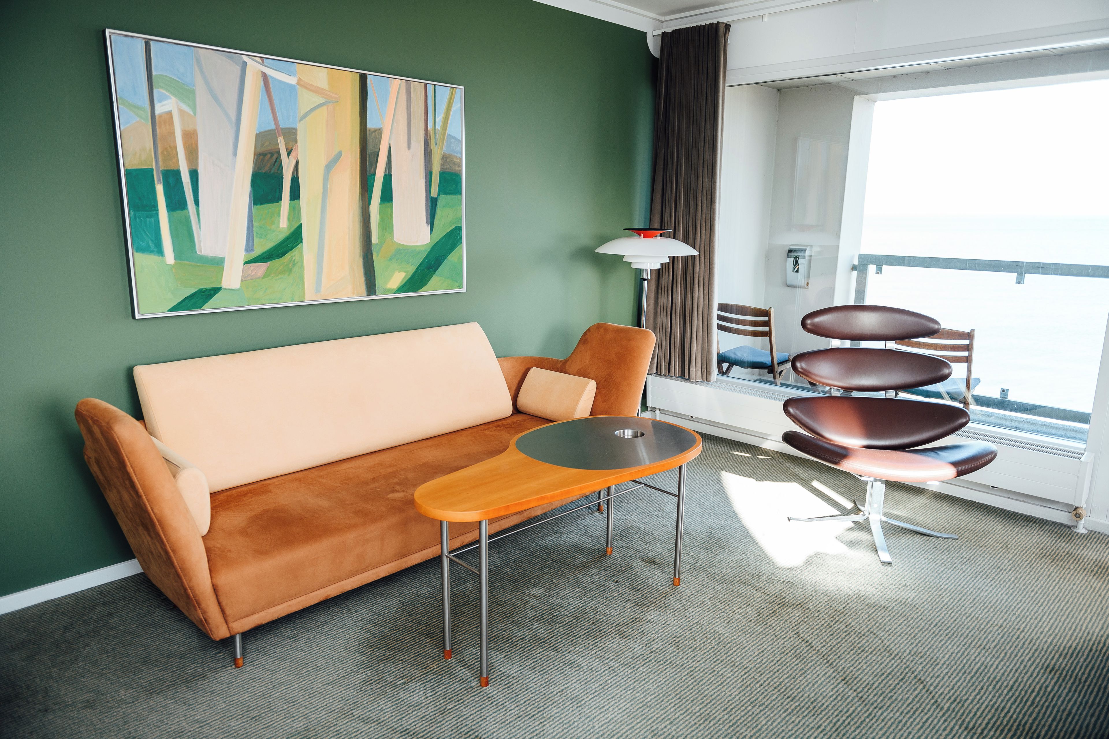 Quarto de hotel moderno com sofá, mesa pequena, cadeira e uma pintura grande na parede.