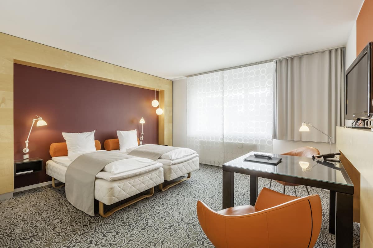 A hotel room with two beds, a desk, a chair, a TV, and a window with curtains.