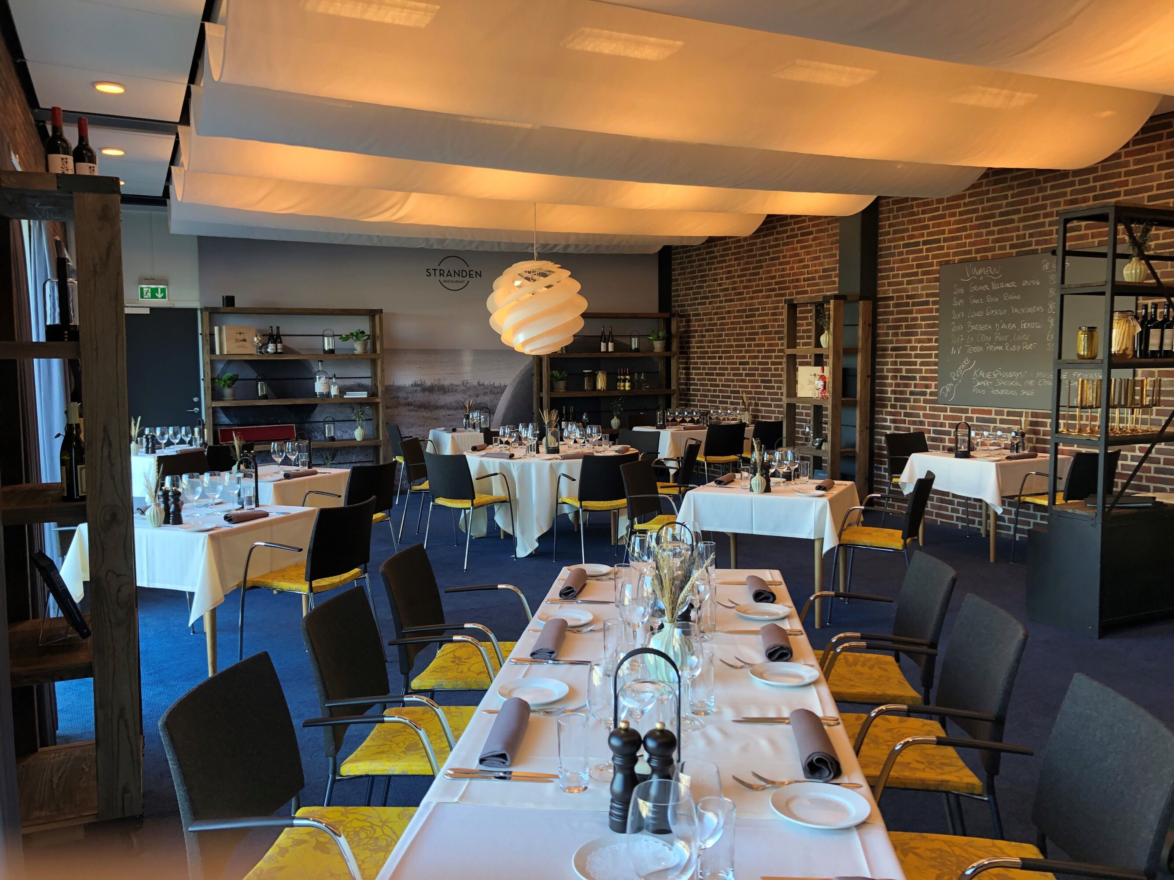 A restaurant with multiple tables set for dining, a brick wall, and shelves with bottles and jars.