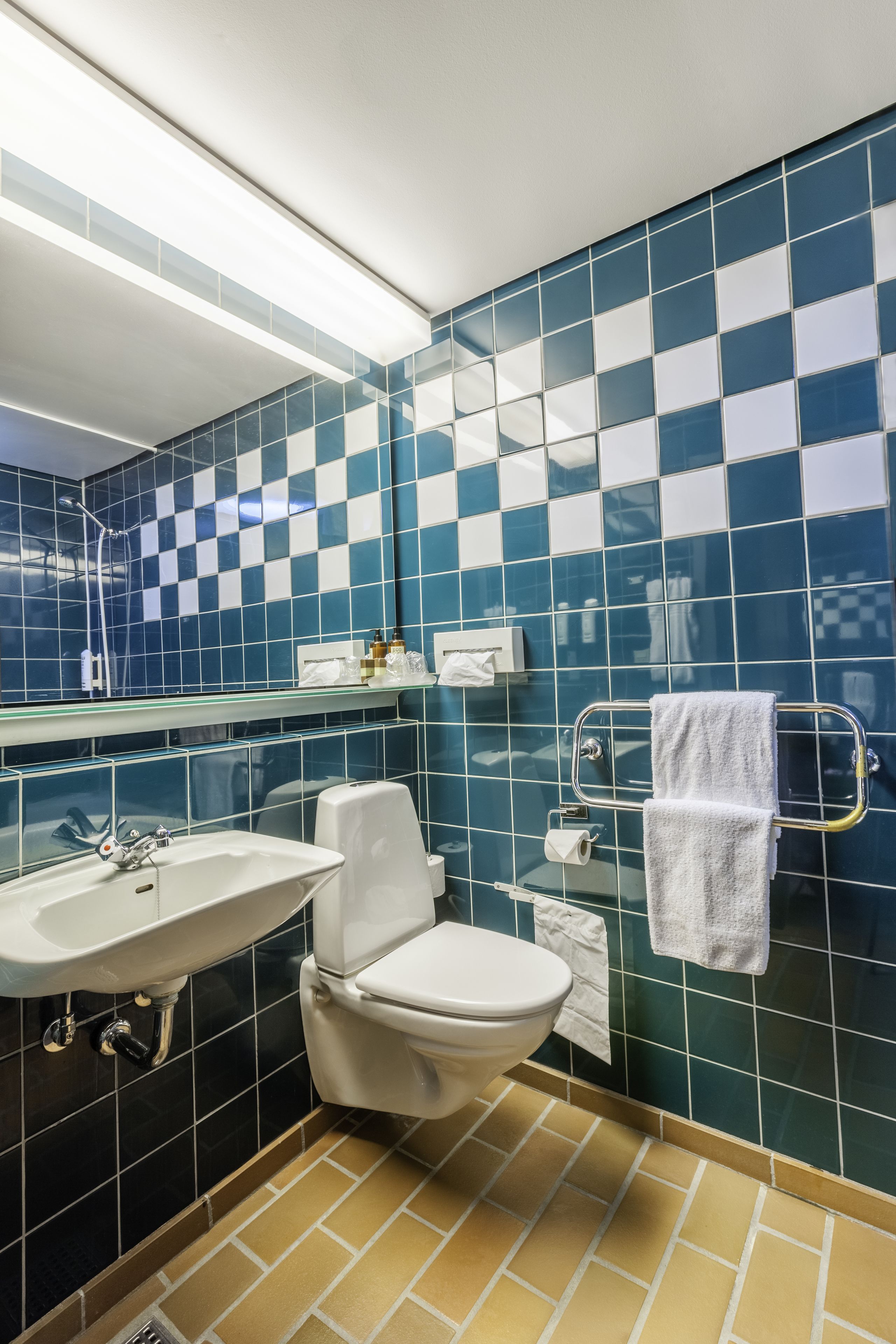 Et badeværelse med en hvid vask, toilet og et spejl. Væggene er fliset med et blåt og hvidt skakmønster.
