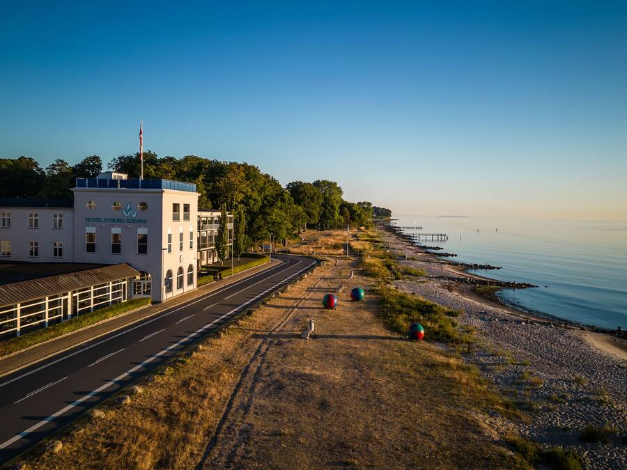 Een kustweg met links het hotel "Hotel Nyborg Strand", een kiezelstrand en een pier die in het water uitloopt, en een heldere lucht.