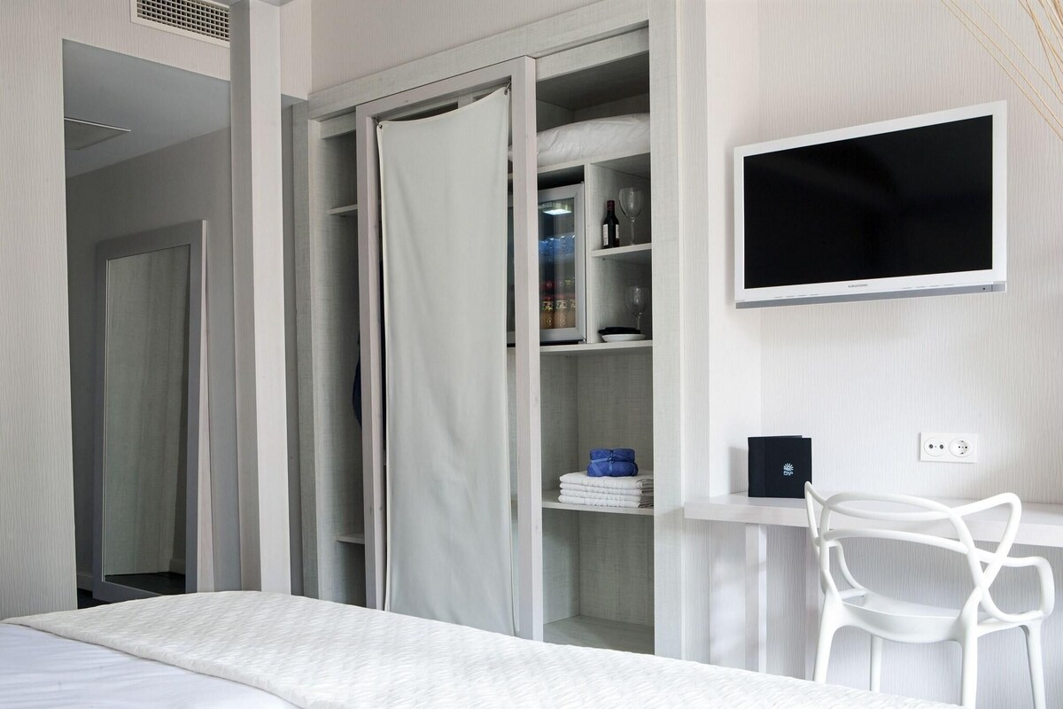 A bedroom with a bed, a white chair, a TV, and a wardrobe with a mirror.