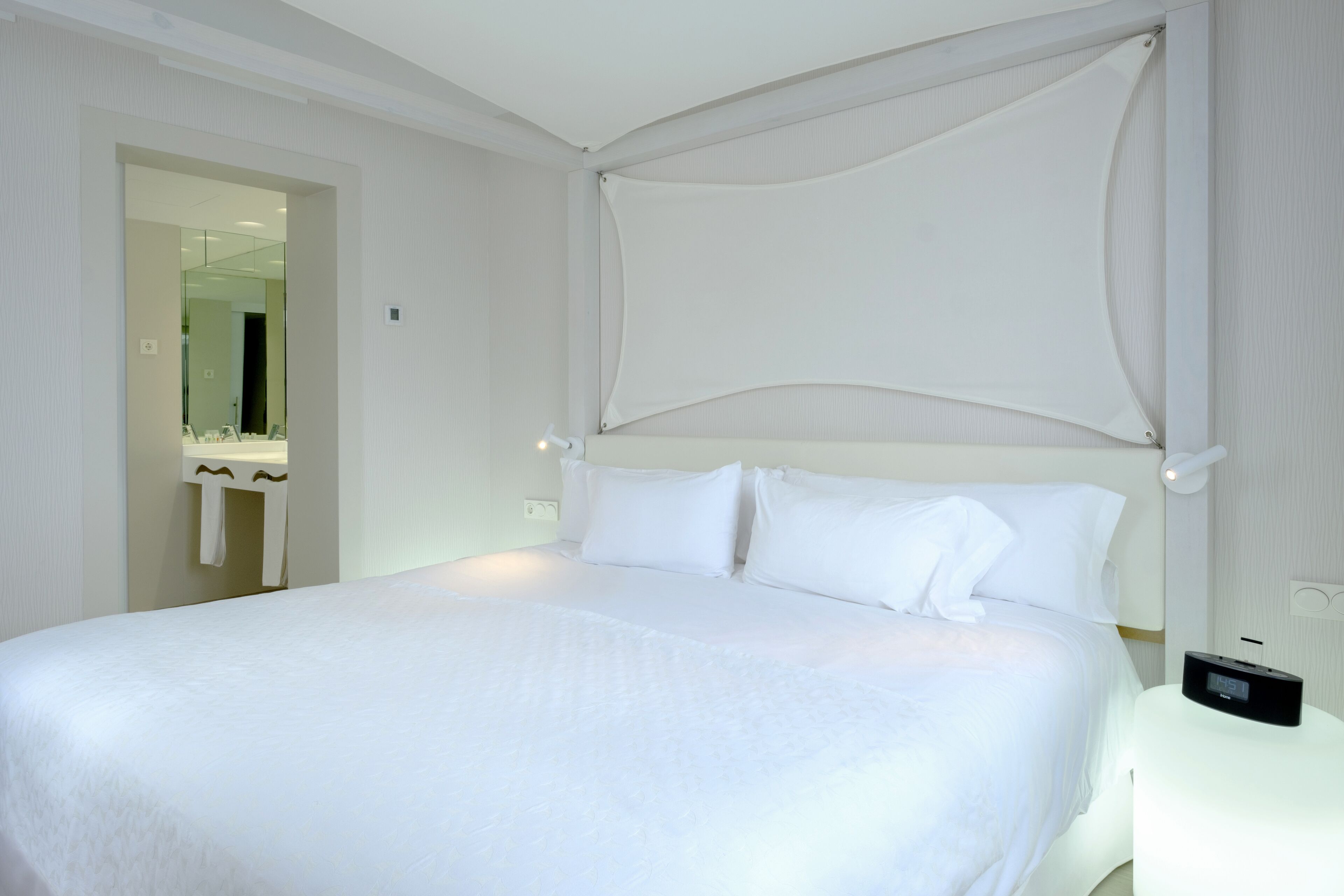 Ein Hotelzimmer mit einem großen Bett, einem Nachttisch mit digitaler Uhr und einem Spiegel in der Ecke.