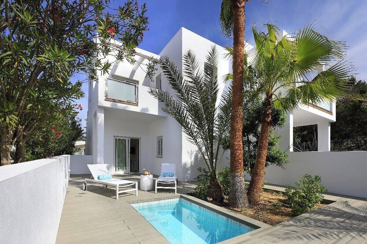 Una villa blanca moderna con piscina, sillas de descanso y palmeras.