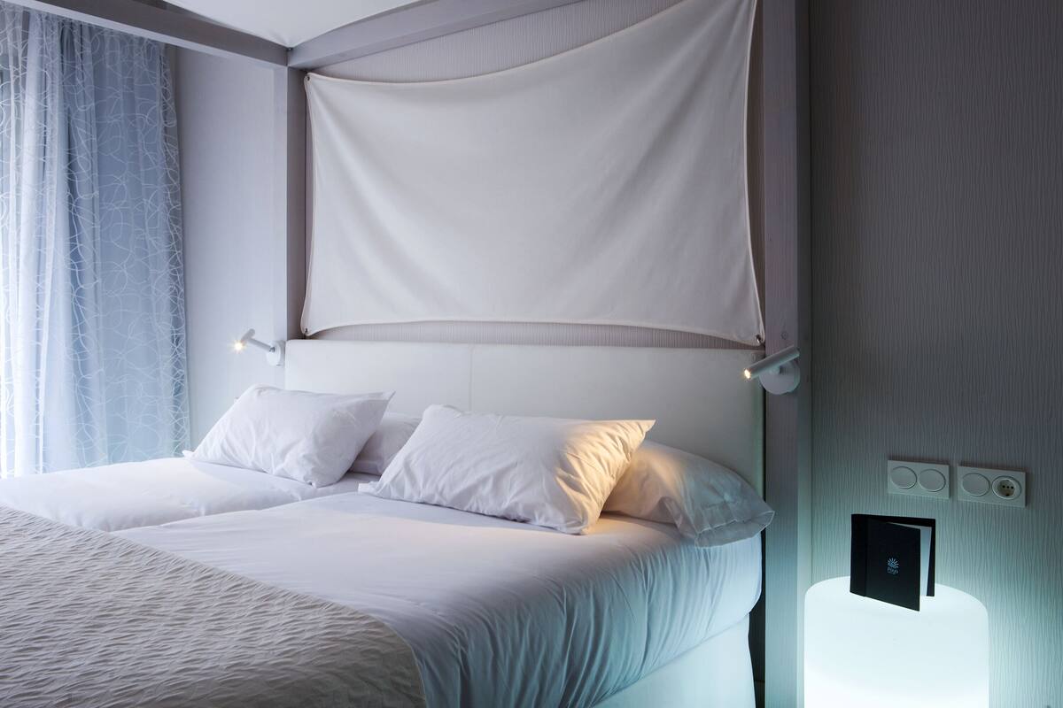 Una cama bien tendida con sábanas blancas y dosel, una mesita de noche con lámpara y un panel de control en la pared.