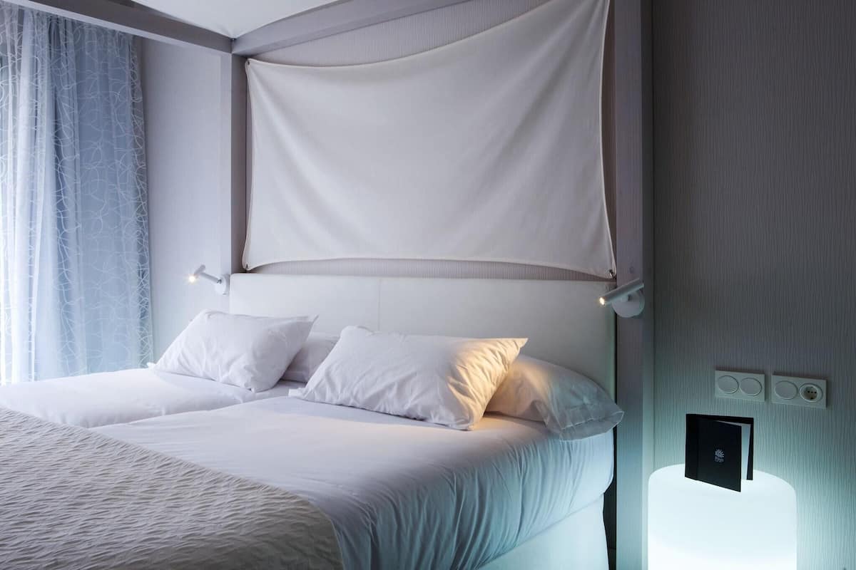 Una habitación de hotel con cama, cortinas blancas, una mesita de noche y una lámpara fijada a la pared.