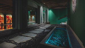 Un espace piscine intérieure paisible, avec des marches en pierre menant à l’eau, un mur végétal et une grande horloge.