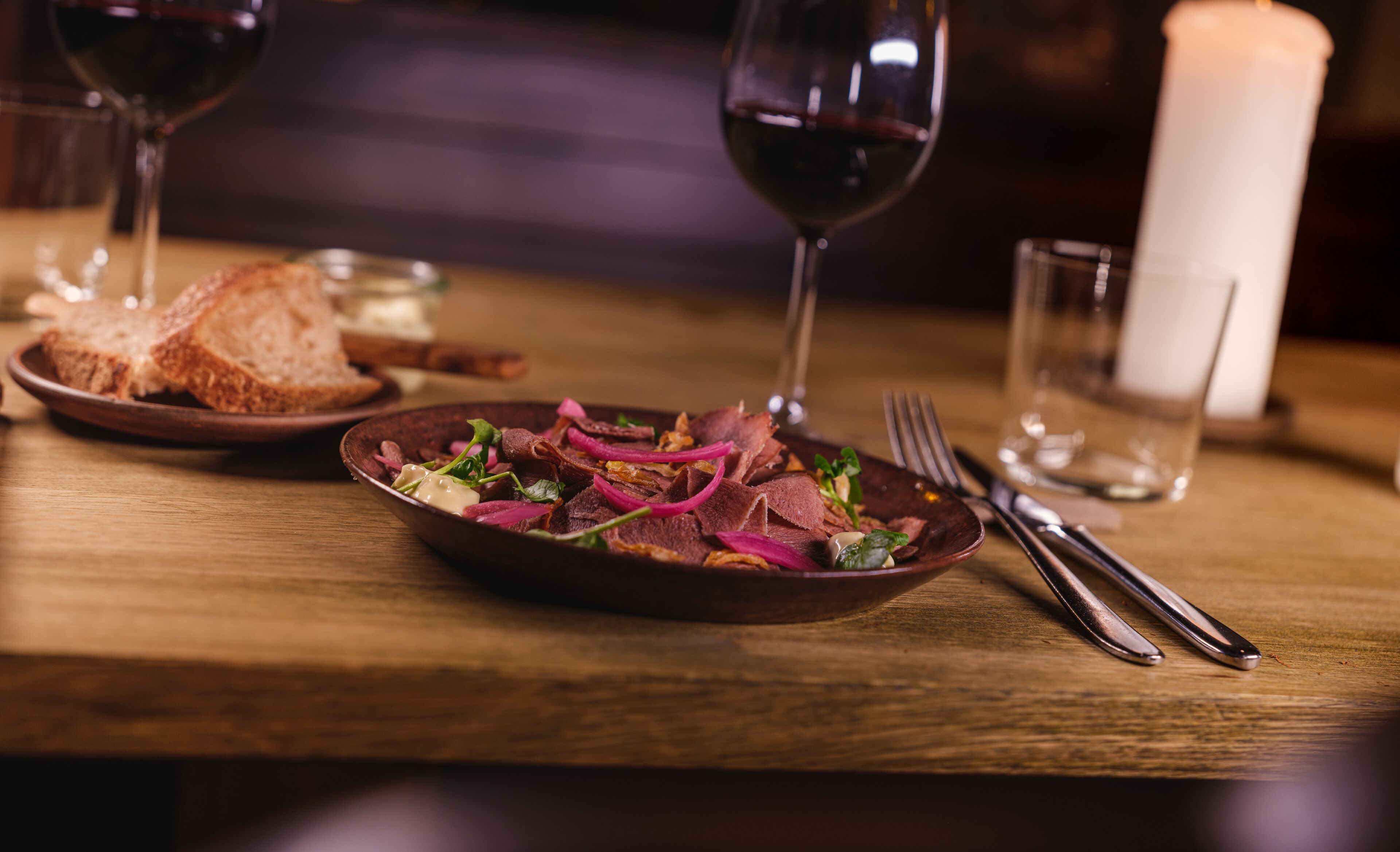 Una mesa de madera con un plato de comida, una copa de vino tinto y una vela.